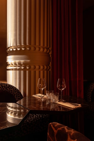 Table élégante près d’une colonne romaine et rideaux rouges, éclairée par une lumière tamisée