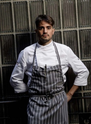 Portrait du chef José Maria Goñi Martínez, en veste et tablier de cuisine, devant un mur de verre cannelé