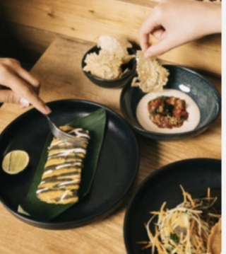 Deux mains dégustent des plats raffinés sur une table en bois, dont un tamal de maïs nappé de sauce et des chips croustillantes avec condiment.
