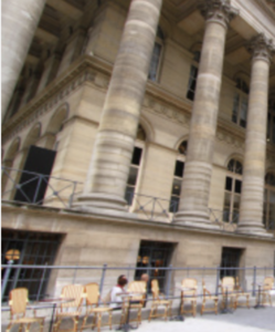 Façade d’un bâtiment historique avec de hautes colonnes classiques, terrasse ensoleillée et quelques clients assis à l’extérieur.