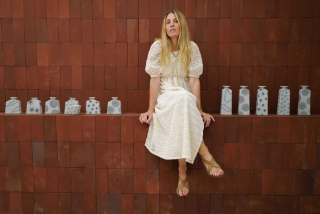 Une femme aux cheveux blonds en robe en dentelle blanche, assise au bord d'un mur en briques rouges, jambes croisées, entourée de vases en porcelaine blanche à motifs pointillés, créant une ambiance élégante et décontractée.
