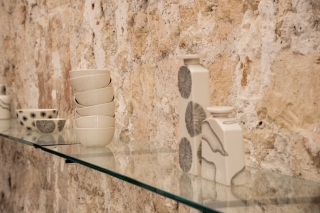 Porcelaine artisanale blanche exposée sur une étagère en verre, incluant des bols empilés et des bouteilles aux motifs abstraits, sur un fond de mur en pierre rugueux, illustrant l'exposition artistique de Saints Pères.