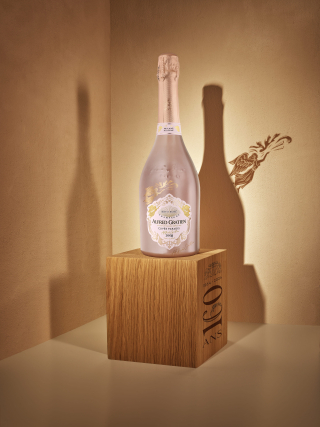 Gros plan d'une bouteille de Champagne Alfred Gratien Cuvée Paradis Brut Rosé millésime 2008 sur un socle en bois commémorant les 160 ans de la Maison, avec une ombre d'ange projetée sur le mur.