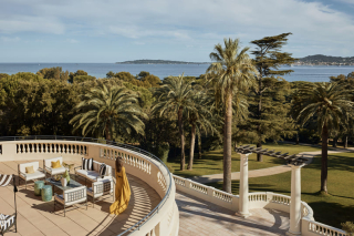 Une femme en robe jaune marchant sur une grande terrasse semi-circulaire de suite, surplombant les jardins de palmiers et la mer au loin.