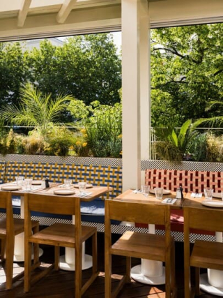 Terrasse de restaurant semi-ouverte entourée de verdure luxuriante, aménagée avec des tables en bois et des banquettes aux motifs tressés colorés.