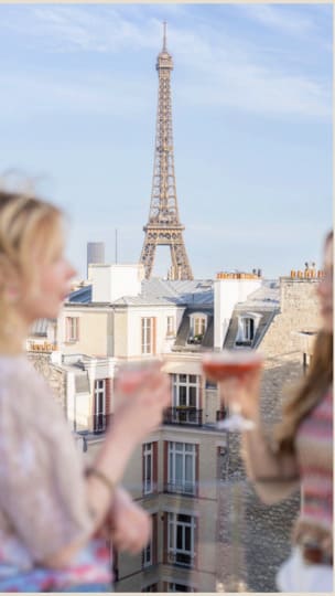 Deux femmes (floues au premier plan) trinquant avec des cocktails sur un rooftop parisien, avec une vue dégagée sur les toits haussmanniens et la Tour Eiffel.