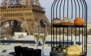 Un présentoir à pâtisseries en forme de cage à oiseaux rempli de mets salés et sucrés, accompagné de deux coupes de champagne, avec la Tour Eiffel s'imposant majestueusement en arrière-plan.