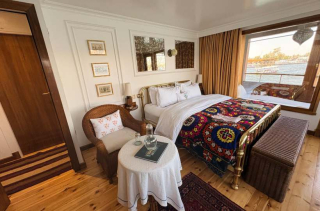 Chambre élégante avec lit double, fauteuil en rotin, petite table, décoration colorée sur le lit et vue sur l’eau par la fenêtre.