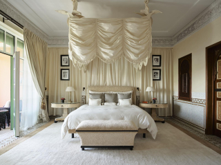 Chambre aux tons ivoire, avec lit surmonté d’un baldaquin drapé à franges, banquette claire, moulures sculptées et portes en bois ouvragé.