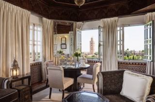 Salon de La Mamounia avec table dressée, fauteuils clairs et fenêtres ouvertes sur les jardins et le minaret de la Koutoubia.